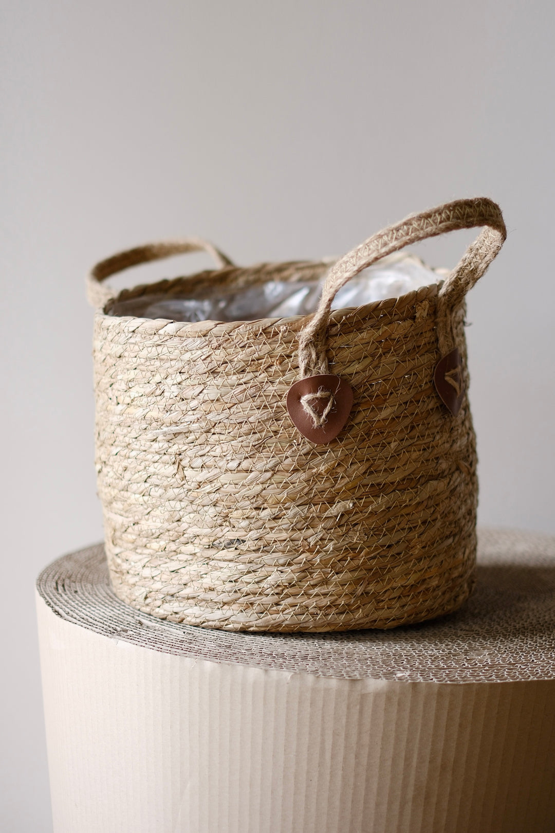 Der Korb Übertopf Alto Ø26 cm von Viva tu Casa, ein geflochtener Korb mit Griffen auf einem strukturierten runden Sockel, kombiniert mühelos rustikalen Charme und modernes Design. Er besteht aus natürlichen Materialien wie Seegras und Baumwolle und ist mit zwei herzförmigen Lederakzenten auf neutralem Hintergrund verziert.