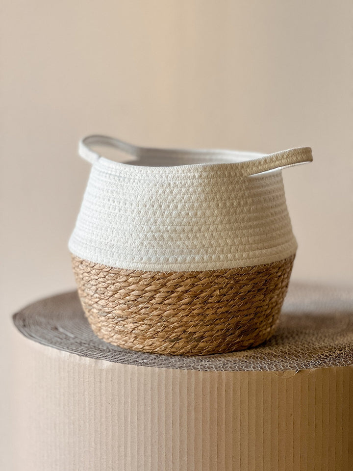 Der Korb Übertopf Halb Weiss Ø27 cm von Viva tu Casa, mit einer Oberseite aus weißem Stoff und einer Unterseite aus gewebtem Seegras, ruht elegant auf einer strukturierten, runden Kartonoberfläche und verkörpert modernes Design.