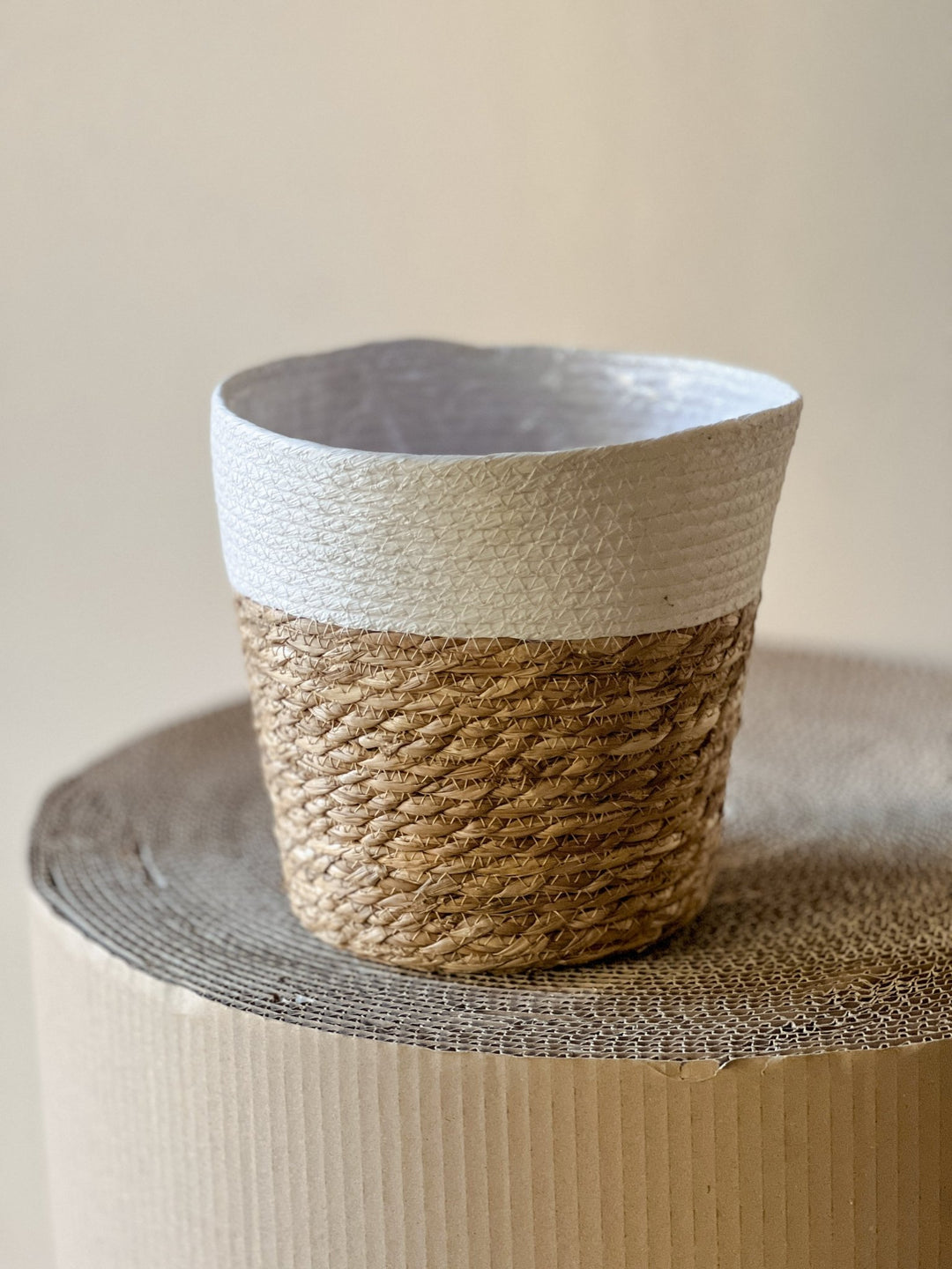 Der Übertopf Korb Halb Weiss Ø24 cm von Viva tu Casa mit seinem Oberteil in Weiß und einem Unterteil, das an einen natürlichen, geflochtenen Pflanzenkorb erinnert, ruht auf einer runden Kartonoberfläche. Dieser kleine Leinentopf hat ein strukturiertes Design, das vor dem neutralen beigen Hintergrund eine rustikale Atmosphäre ausstrahlt.