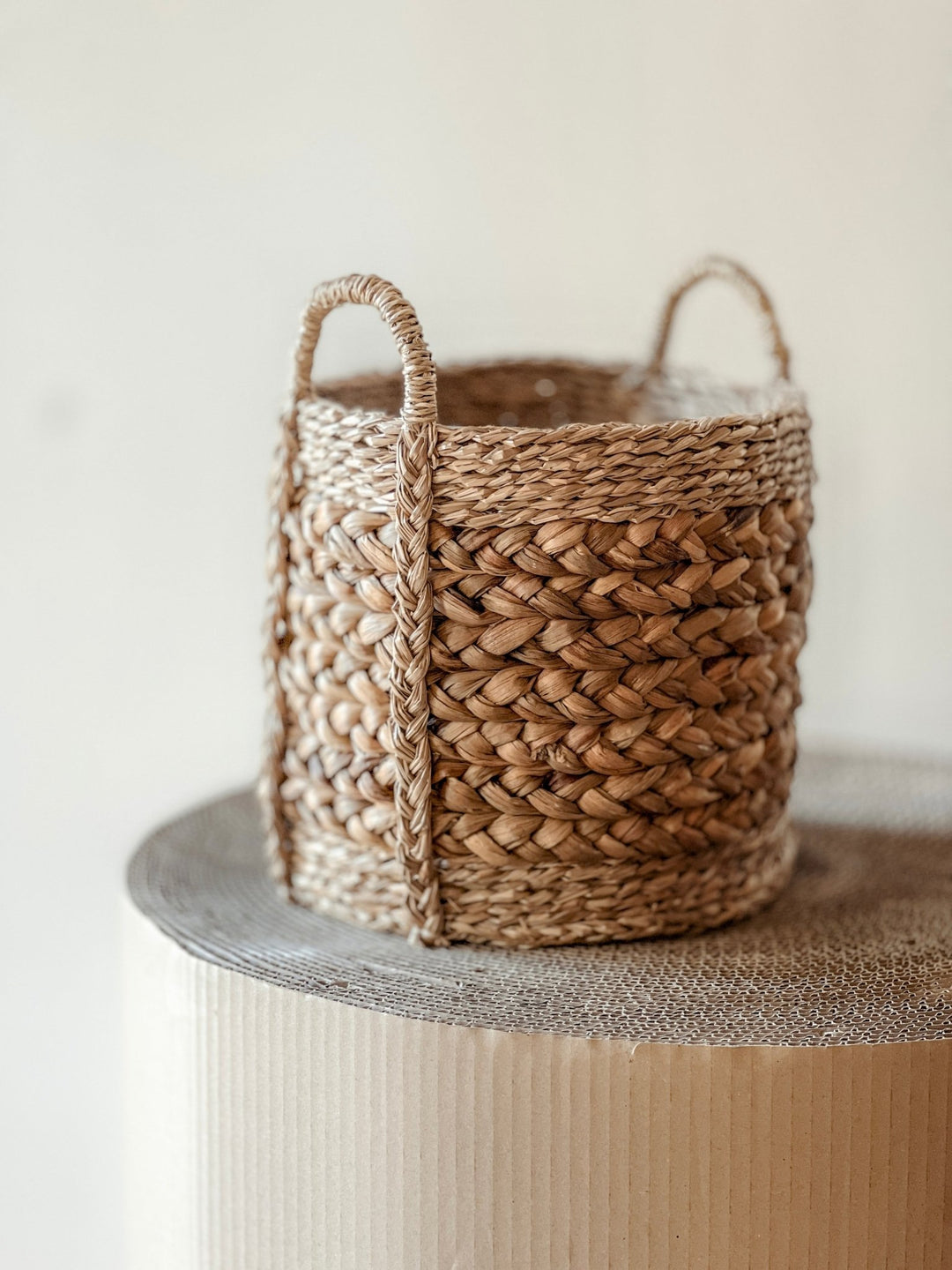 Ein geflochtener Weidenkorb mit Griffen liegt auf einer strukturierten Oberfläche. Dieser zylindrische Übertopf Korb Nature Ø26 cm von Viva tu Casa besticht durch ein kompliziertes Flechtmuster, das rustikalen Charme verleiht. Der aus Materialien wie Seegras gefertigte, sanft verschwommene Hintergrund betont die exquisiten Details des Korbs.