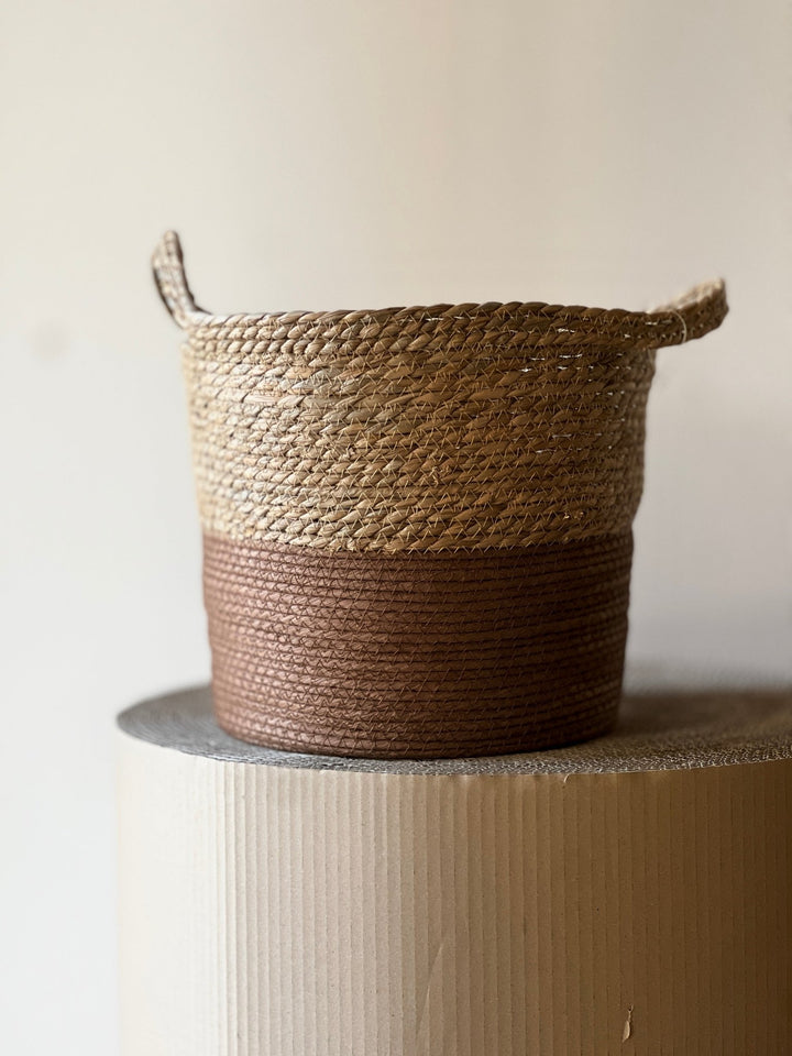Der Übertopf Braun Ø26 cm von Viva tu Casa ist ein geflochtener Seegraskorb mit zweifarbigem Design. Er besteht aus einer naturbeigen oberen Hälfte und einer braunen unteren Hälfte, komplett mit zwei kleinen Griffen. Auf einem strukturierten, runden Sockel präsentiert, unterstreicht der neutrale Hintergrund den Charme dieses Pflanzenkorbs.