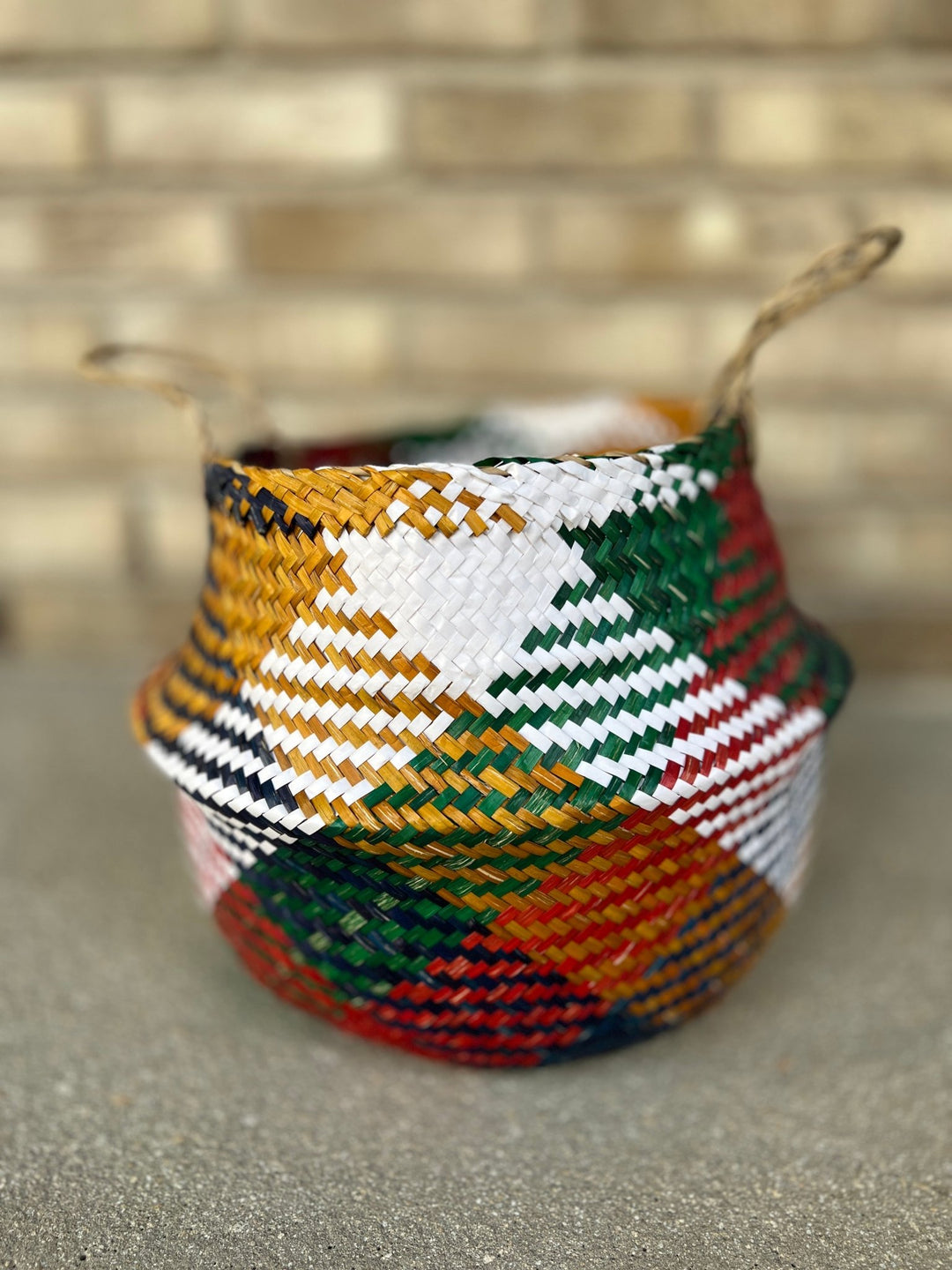 Ein lebhafter Übertopfkorb Farbig Ø26 cm von Viva tu Casa, verziert mit komplizierten Mustern in Gelb, Weiß, Grün, Rot und Schwarz, steht auf einer grauen Oberfläche. Der Korb betont rustikale Eleganz und verfügt über zwei Seilgriffe vor einem strukturierten Ziegelwandhintergrund.