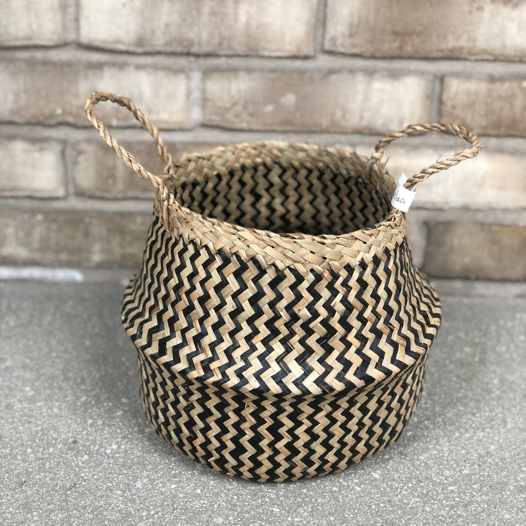 Der Übertopf Korb - Faltkorb Ø18 cm von Viva tu Casa, ein stilvolles Accessoire, zeigt ein Zickzack-Muster in Schwarz und Naturfasern, das sich perfekt von einer Betonoberfläche mit Ziegelmauerhintergrund abhebt.