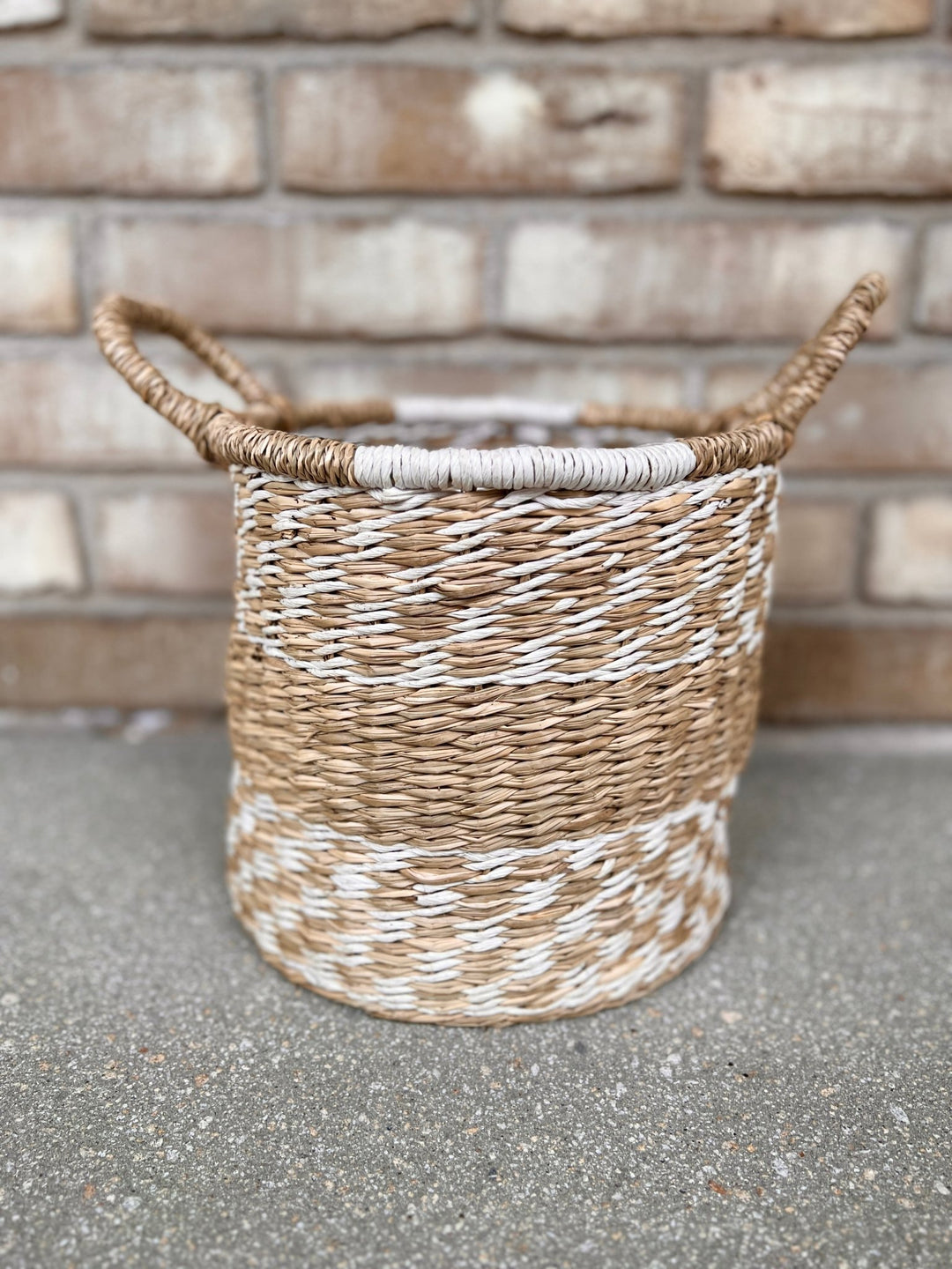 Ein geflochtener Übertopf Korb Natur Ø26 cm von Viva tu Casa mit zwei Griffen steht auf einer Steinfläche vor einer Backsteinmauer. Der Korb aus Seegras weist abwechselnd helle und dunkle Flechtmuster auf.