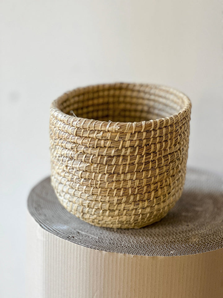 Ein geflochtener, runder Korb von Viva tu Casa strahlt rustikale Eleganz aus, da er auf einer strukturierten, zylindrischen Oberfläche ruht. Der Übertopf Korb - Natur Ø29 cm ist aus Naturfasern gefertigt und weist ein dicht gewebtes Muster mit abwechselnd hellen und dunklen Farbtönen auf. Der Hintergrund bleibt schlicht und neutral.