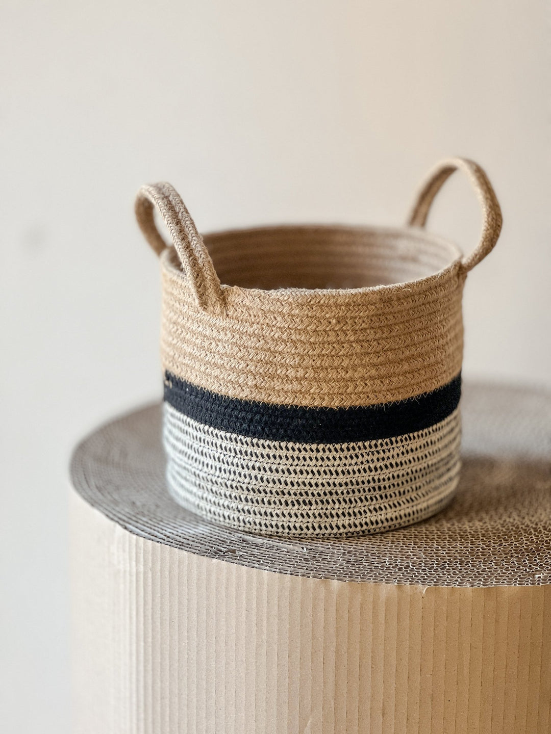 Der Übertopf Korb Streif schwarz Ø23 cm von Viva tu Casa steht auf einer gewellten Oberfläche. Dieser geflochtene Korb, perfekt als Pflanzenkorb, zeigt eine natürliche Beigefarbe mit schwarzen Streifen und hat einen Boden aus weißer Baumwolle. Der Hintergrund ist weich und neutral im Ton.